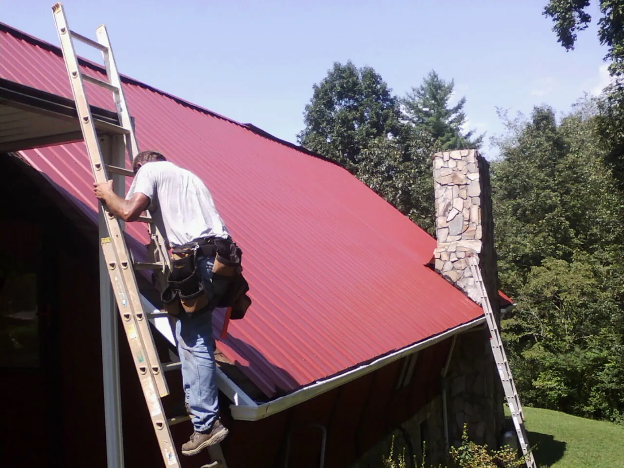Roofer on a ladder beside a red metal roof and stone chimney — Soffit & Fascia Repair by Roofing Brothers in Owings Mills
